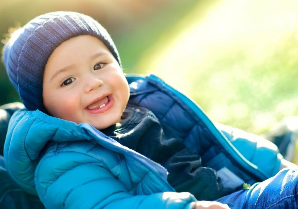 Portrait of a cute little boy enjoying spring sunny day, wearing blue jacket and hat sitting on the grass field, happy day outdoors